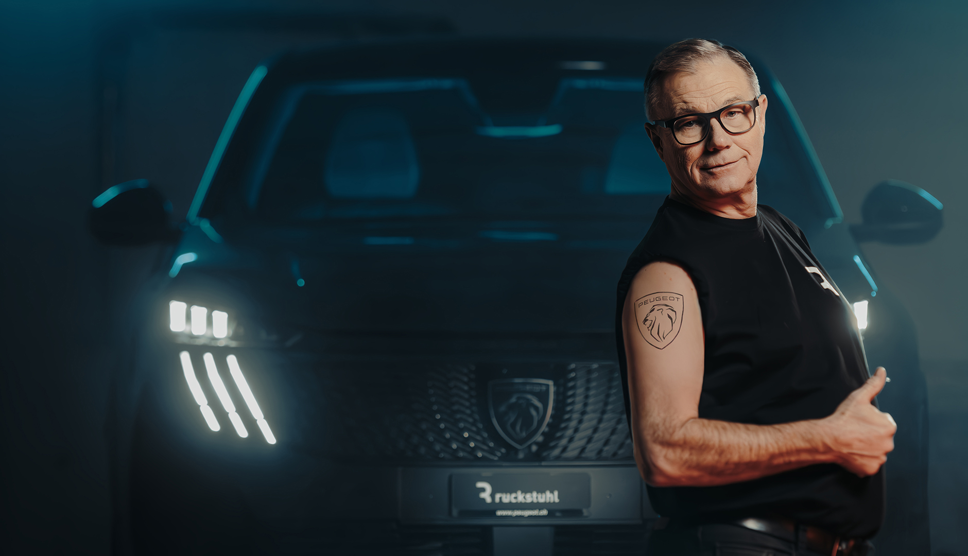 Mann mit Peugeot-Logo-Tattoo steht vor beleuchtetem Auto.