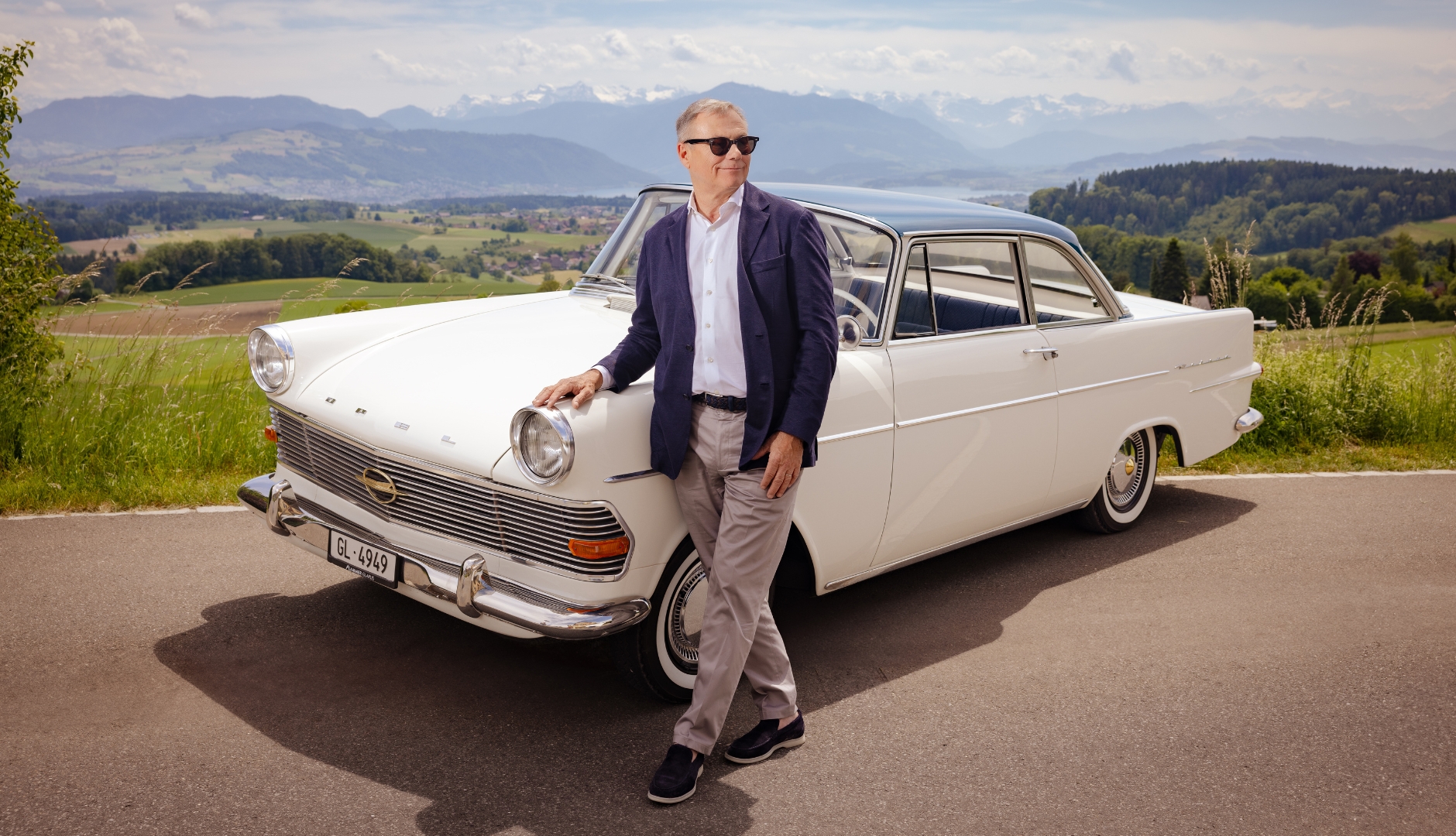 Mann lehnt an einem klassischen weissen Auto mit idyllischer Landschaft im Hintergrund.